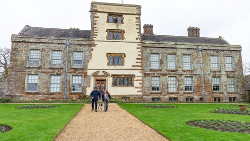 Two visitors and a dog walking in the formal gardens at Canons Ashby in winter with the house behind